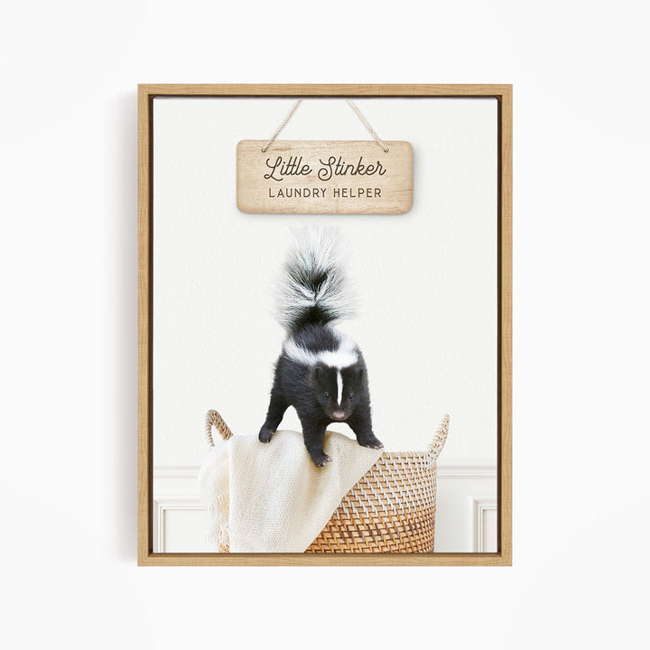 A framed image of a black and white skunk standing on a woven basket, with a wooden sign above it that reads "Little Stinker Laundry Helper".