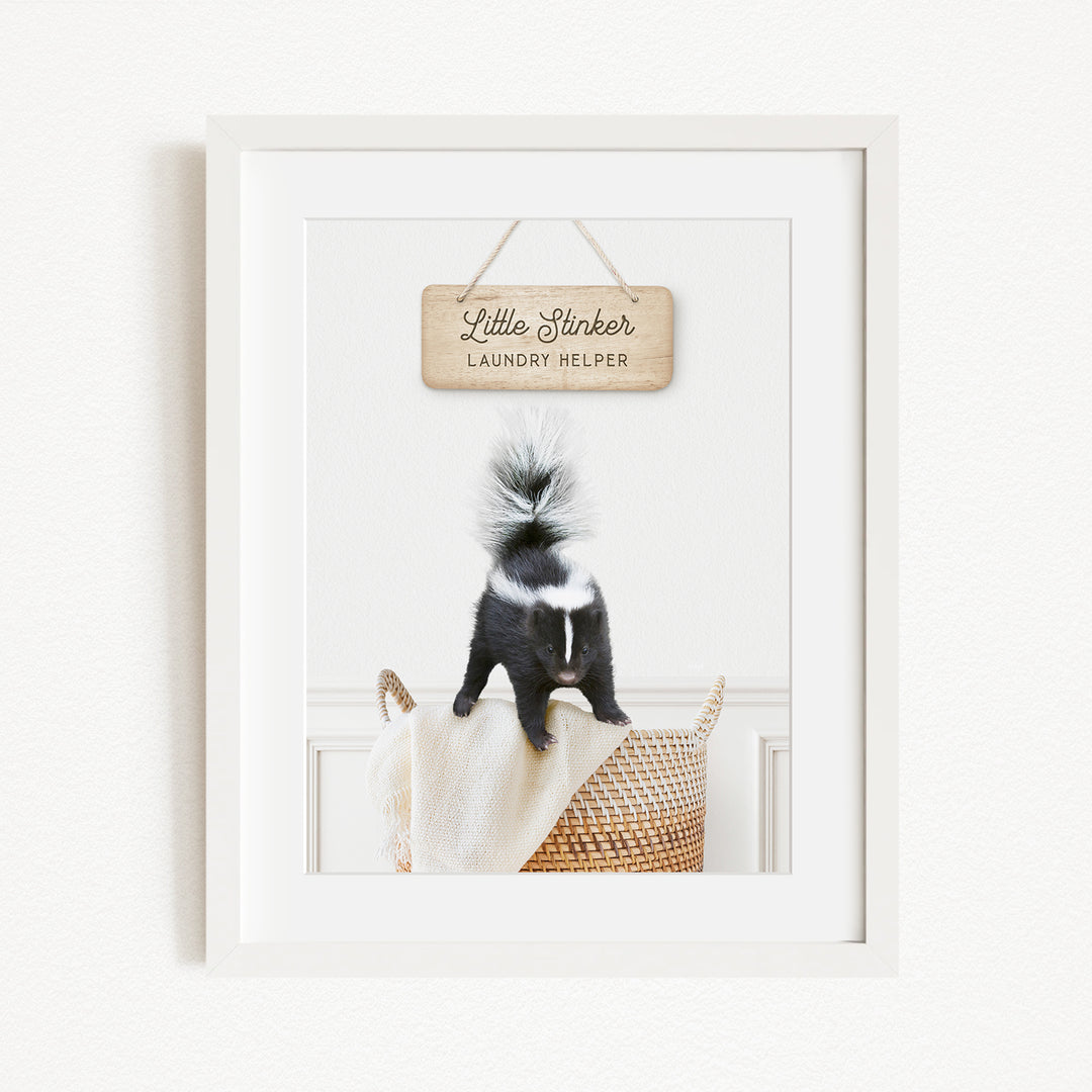 A framed image of a black and white skunk standing on a woven basket, with a wooden sign above it that reads "Little Skunk Laundry Helper".