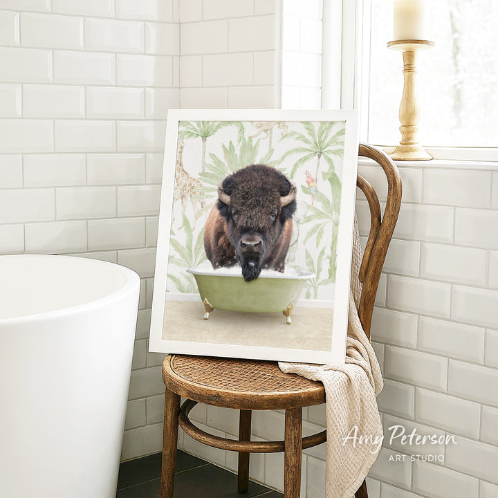A framed image of a bison resting in a bathtub, surrounded by tropical foliage, is displayed on a wooden chair in a bathroom setting.
