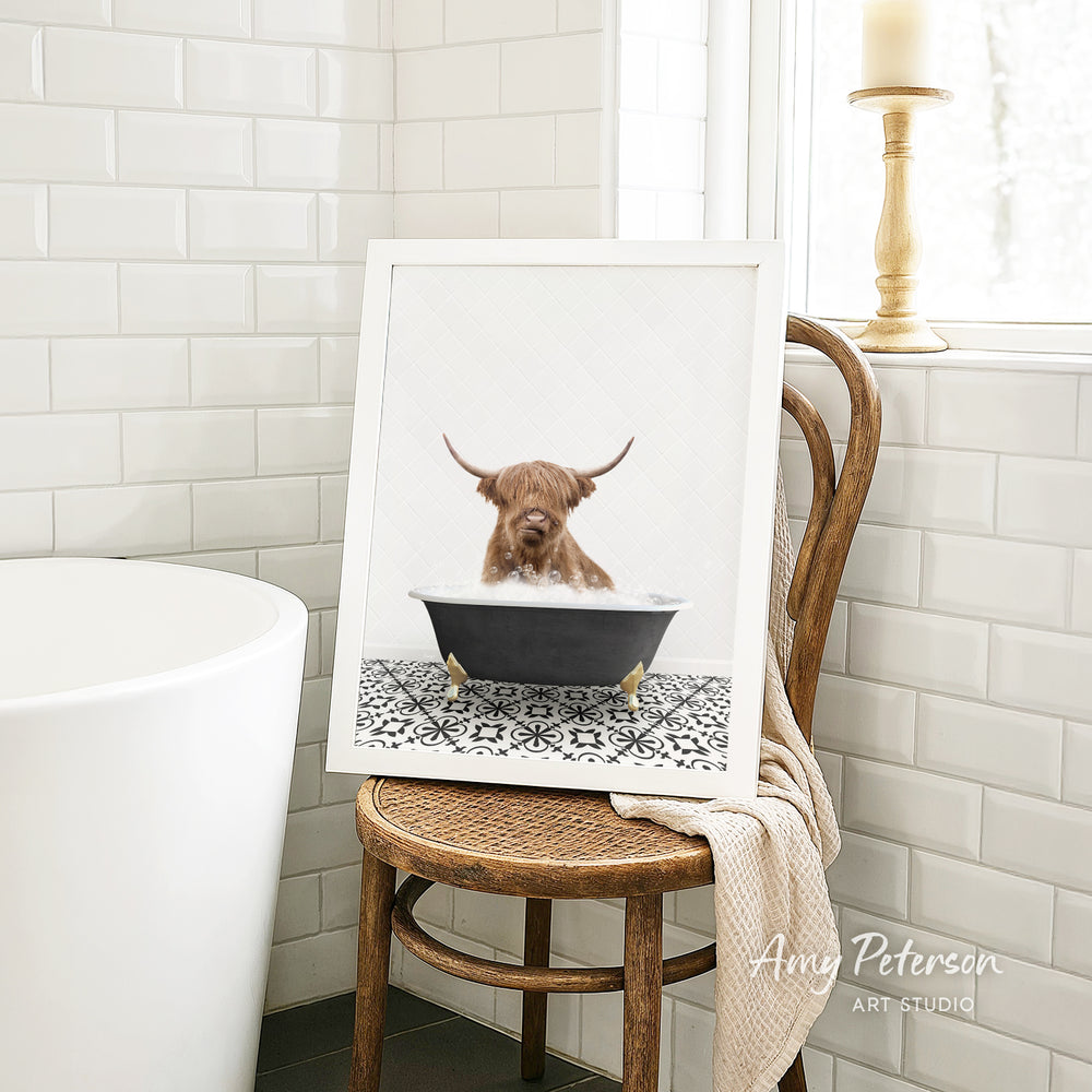 A framed image of a Highland cow sitting in a bathtub, with a towel draped over the back of a wooden chair in a bathroom setting.