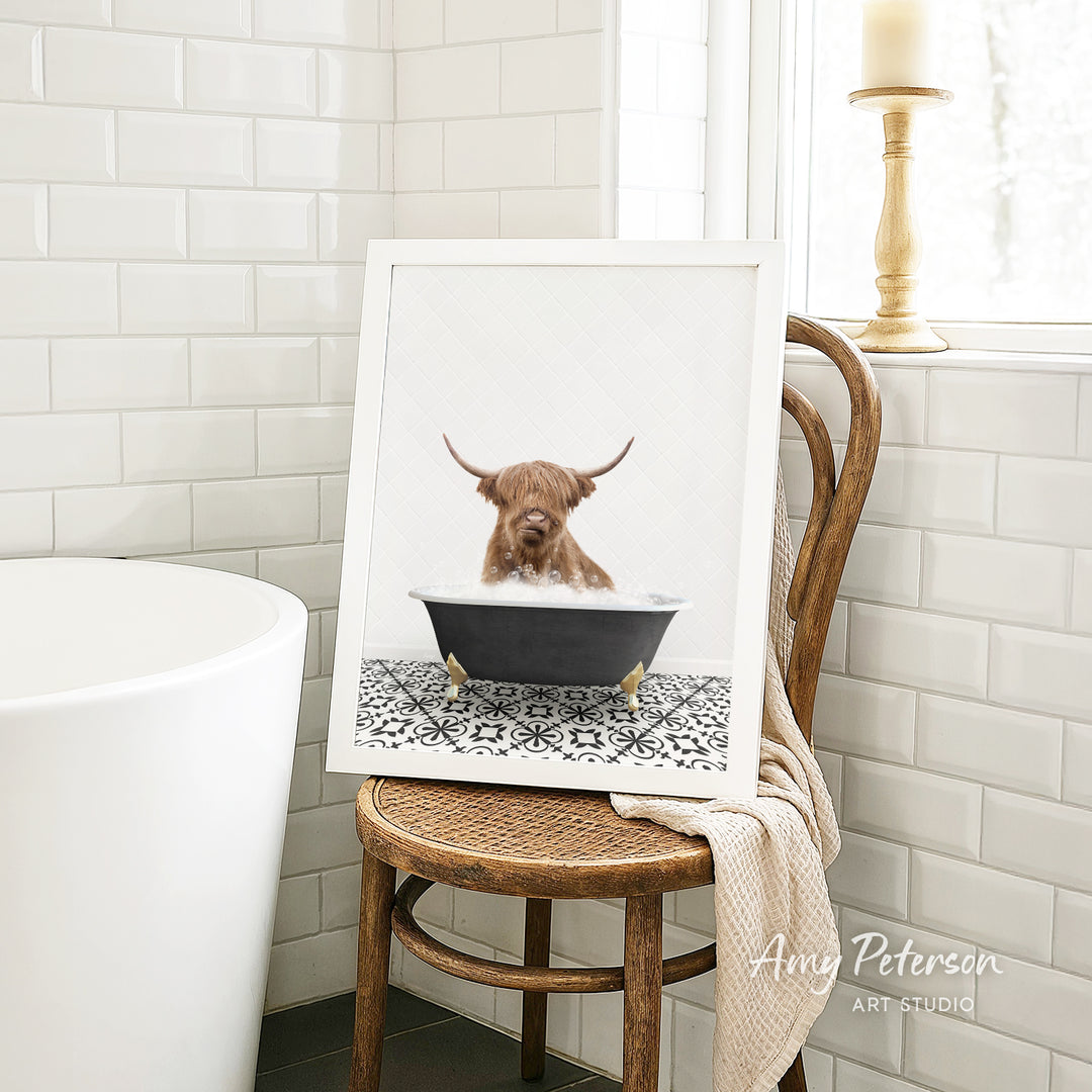 A framed image of a Highland cow sitting in a bathtub, with a towel draped over the back of a wooden chair in a bathroom setting.
