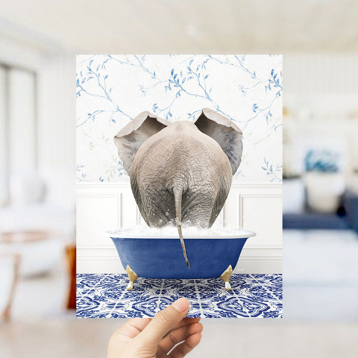 A hand holds up a framed image of an elephant standing in a blue bathtub filled with water, with a patterned tile floor in the background.