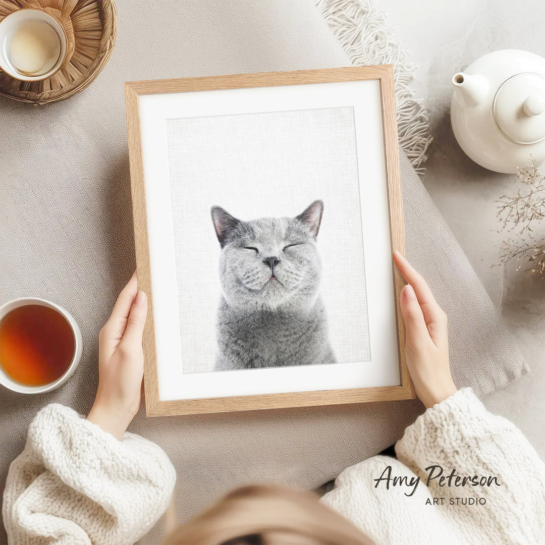 A person is holding a framed picture of a smiling gray cat, with a teapot, a cup of tea, and a bowl of tea visible in the background.