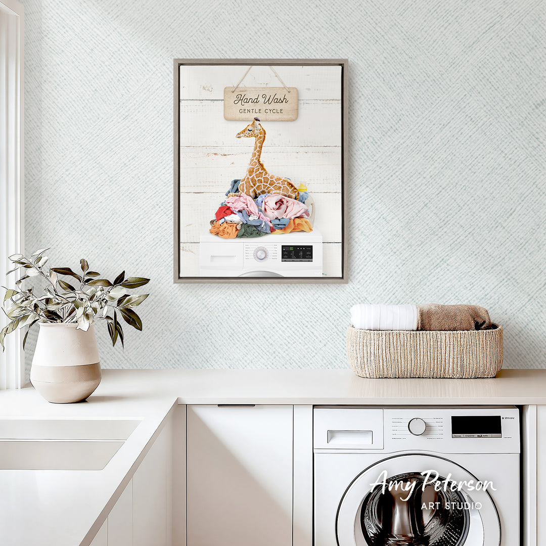 a framed illustration of a giraffe on a pile of laundry, with a sign above it that reads "Hard Work Gentle Cycle". The scene is set in a kitchen or laundry room with a washing machine and a basket of folded clothes.