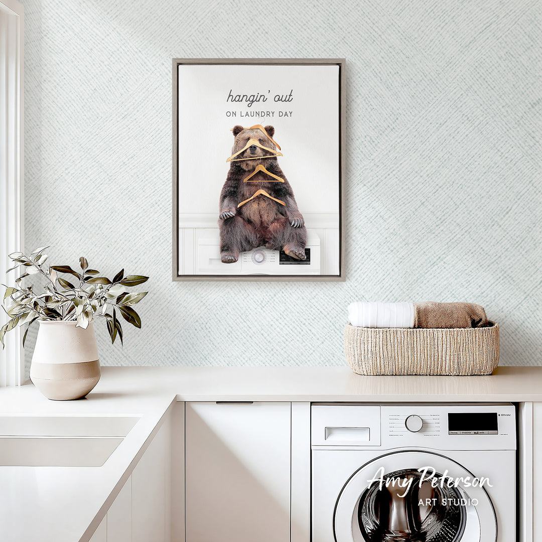 a framed poster of a bear sitting on a shelf, with the text "hangin' out on laundry day" written above it. The poster is displayed on a wall above a white countertop, which has a basket of towels and a potted plant on it. There is also a washing machine visible in the background.