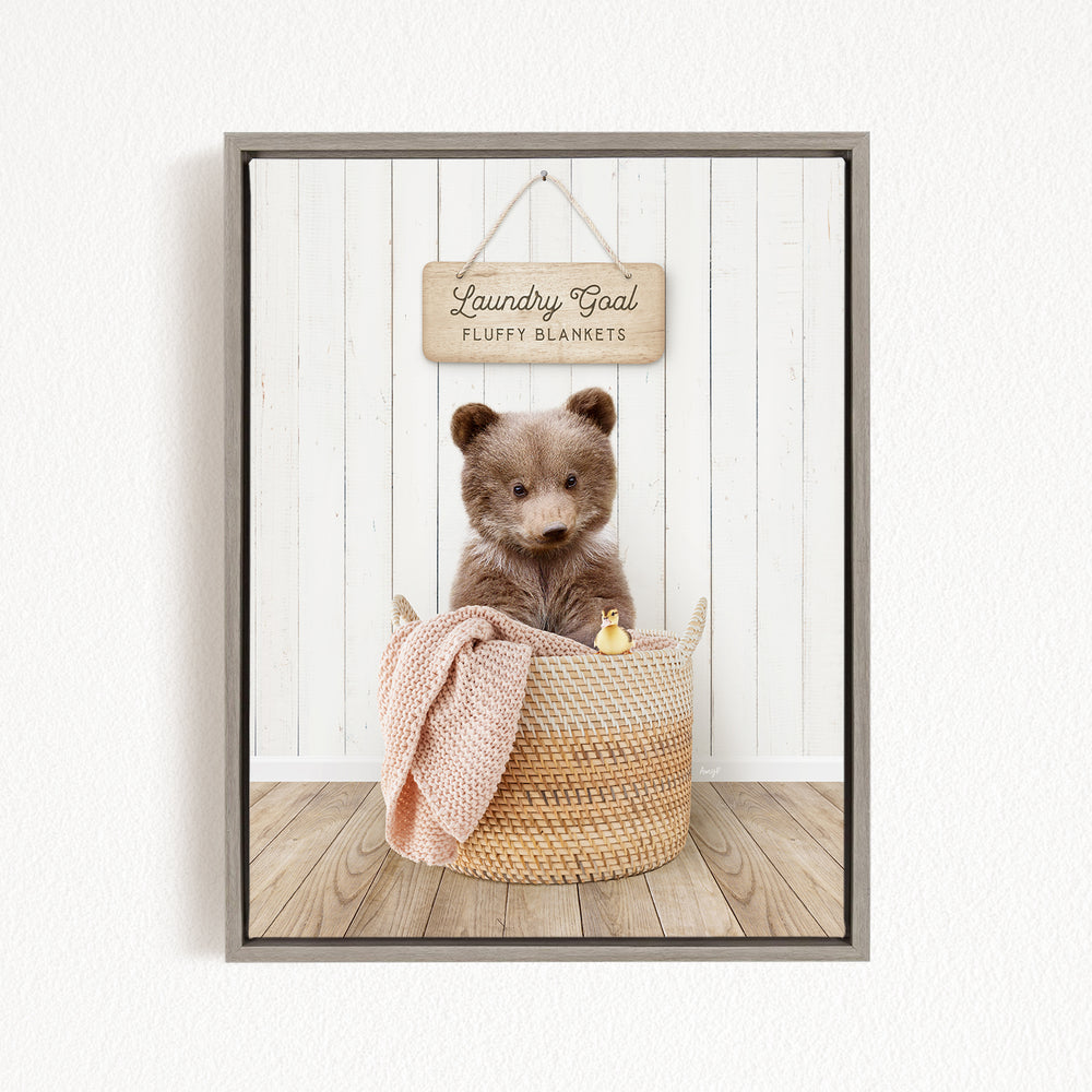 A brown teddy bear sits in a woven basket filled with blankets, with a wooden sign above that reads "Laundry Goal Fluffy Blankets".