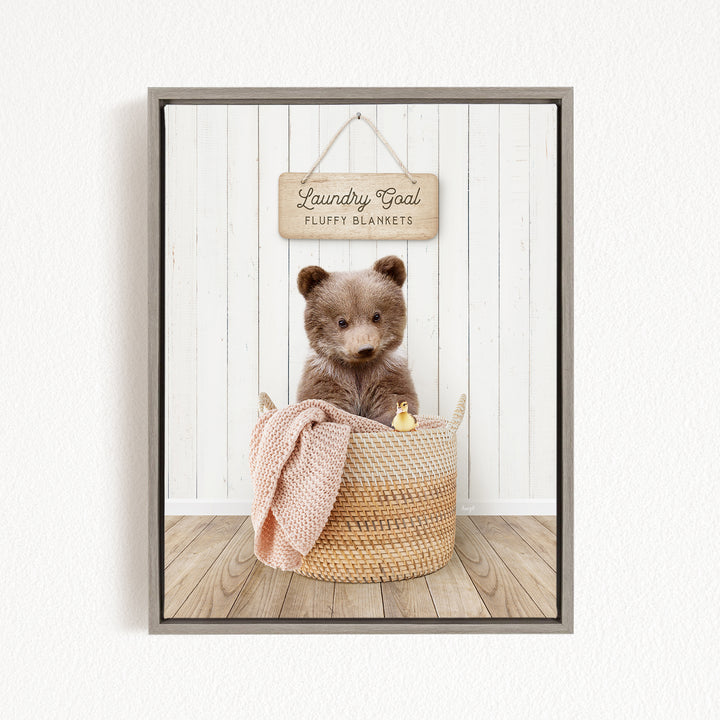 A brown teddy bear sits in a woven basket filled with blankets, with a wooden sign above that reads "Laundry Goal Fluffy Blankets".
