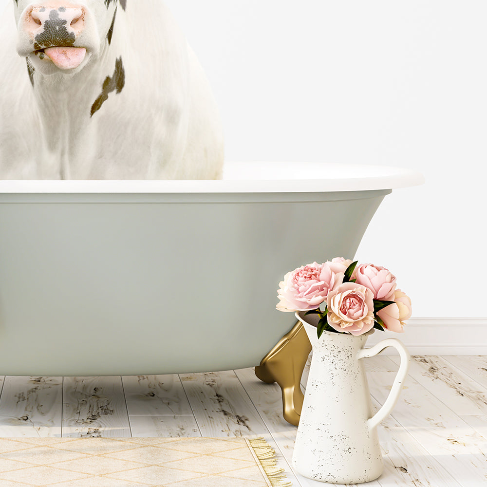 A white cow with black spots is sitting in a white bathtub, with a vase of pink flowers next to it on a wooden floor.