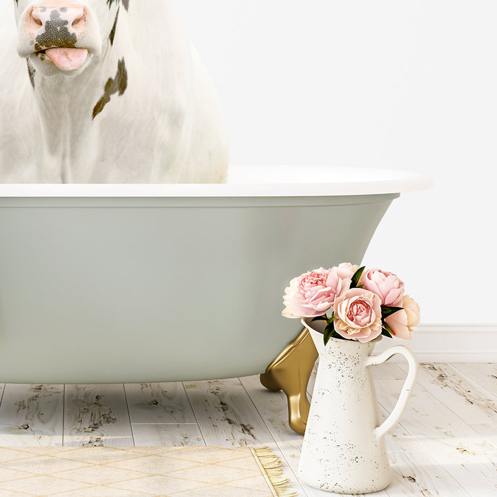 A white cow with black spots is sitting in a white bathtub, with a vase of pink flowers next to it on a wooden floor.