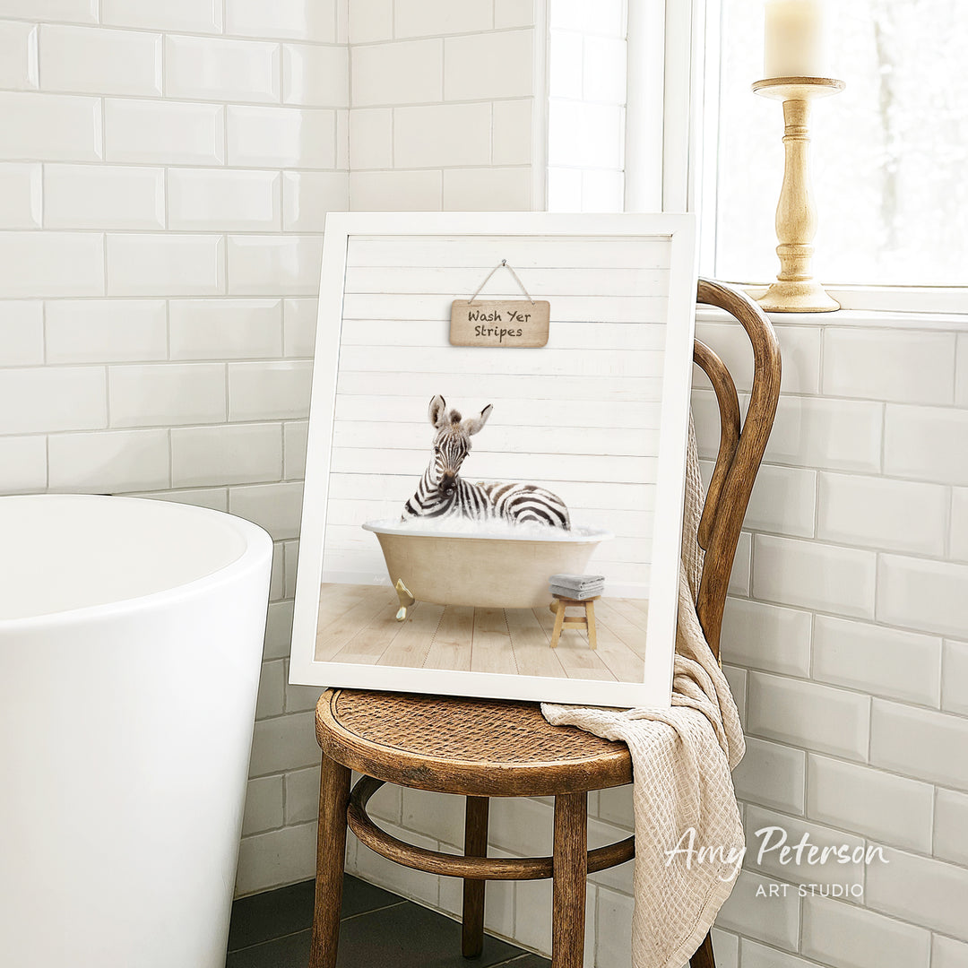 A framed print of a zebra sitting in a bathtub is displayed on a wooden chair in a bathroom setting.