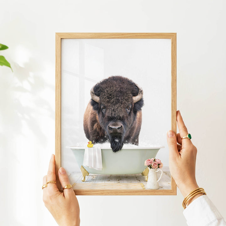 A person's hands are holding a framed picture of a buffalo in a bathtub, with a vase of flowers and a towel nearby.