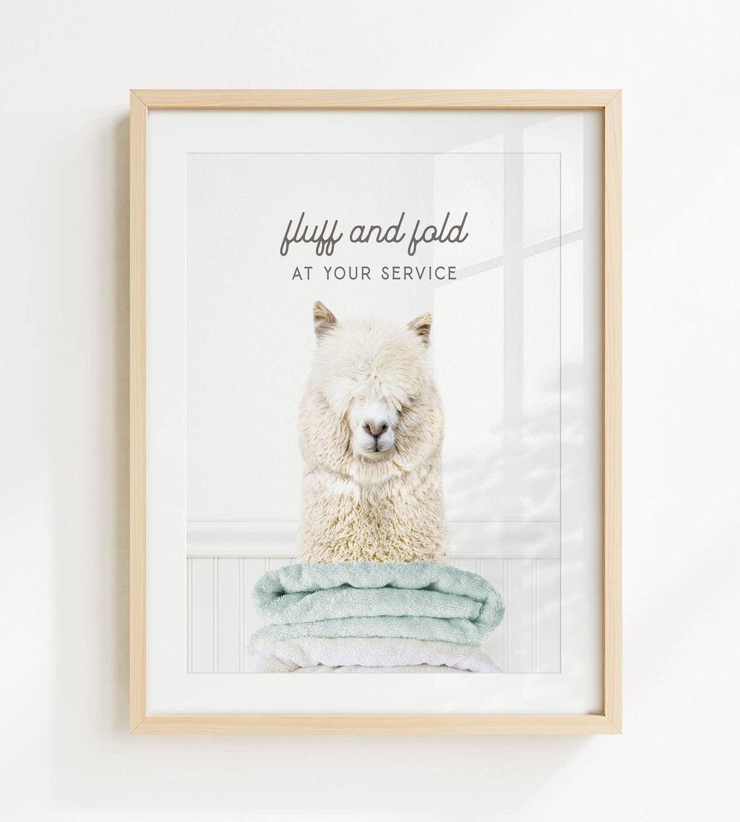 A framed image of a fluffy white alpaca sitting on a stack of light blue towels, with the text "fluffy and folded at your service" written above the image.
