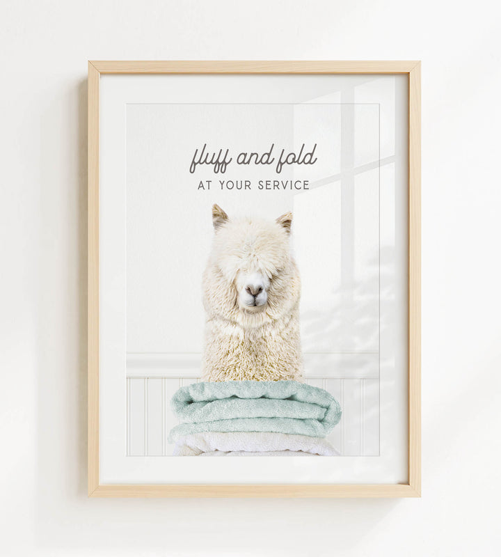 A framed image of a fluffy white alpaca sitting on a stack of light blue towels, with the text "fluffy and folded at your service" written above the image.