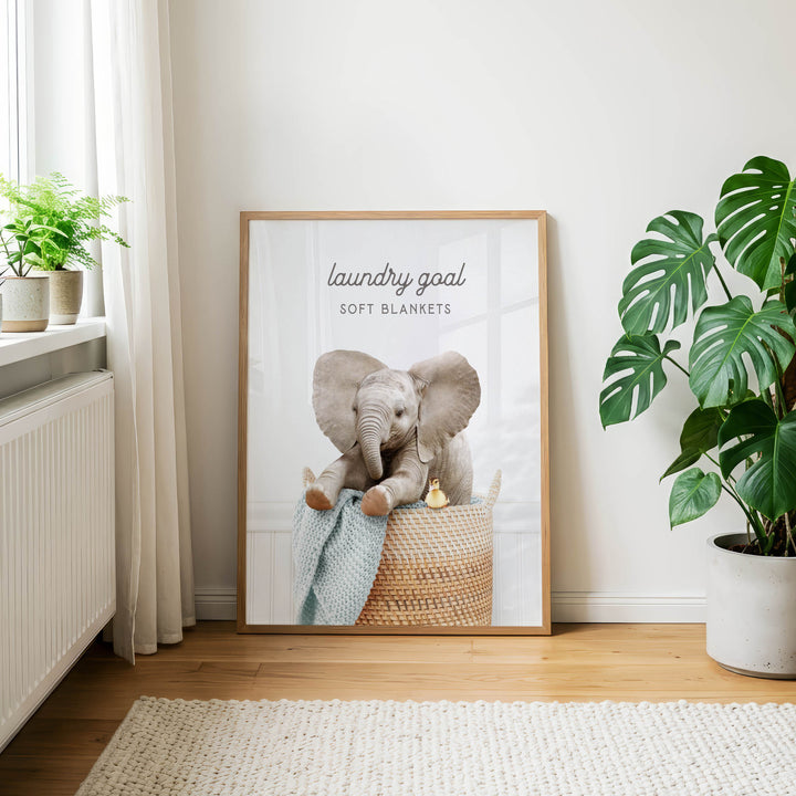 A framed poster of an elephant sitting in a basket, with the text "laundry goal soft blankets" on it, is displayed on a wall in a room with a rug and potted plants.