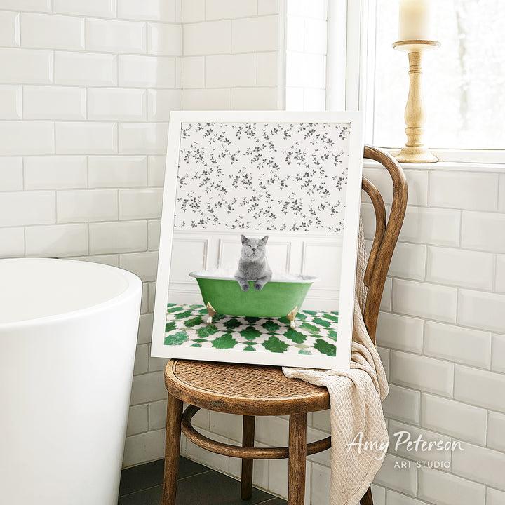 A framed image of a gray cat sitting in a green bathtub, with a floral pattern on the tub, is displayed on a wooden chair in a bathroom setting.