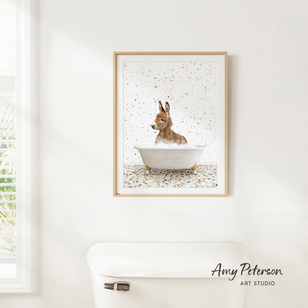 A framed print of a donkey sitting in a bathtub is hanging on a white wall.