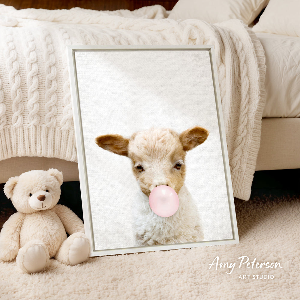 A framed image of a lamb blowing a bubble gum bubble is displayed on a bed, accompanied by a teddy bear.