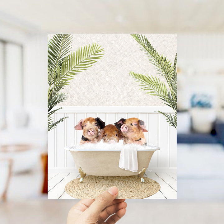 A hand holds up a framed image depicting three adorable pigs sitting in a bathtub filled with bubbles, surrounded by lush green palm leaves.