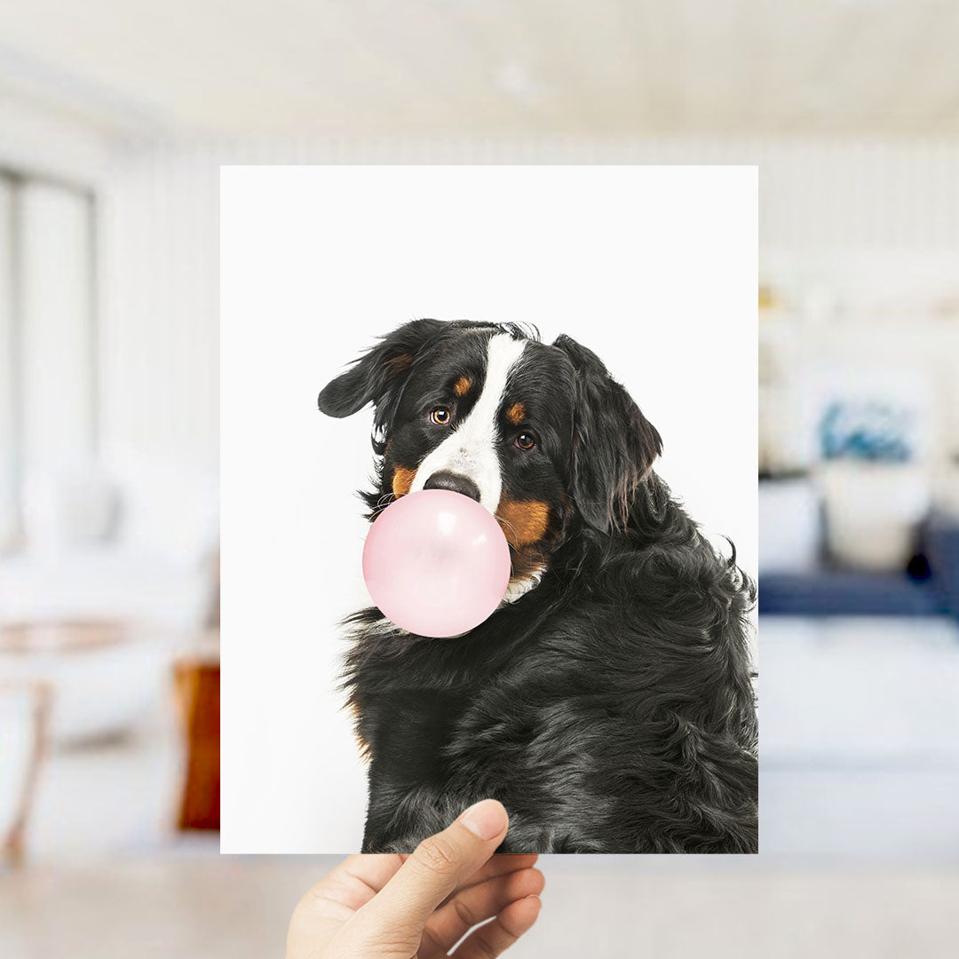 A black and brown dog is blowing a bubble with a pink bubble gum bubble, as a person holds up the photo.