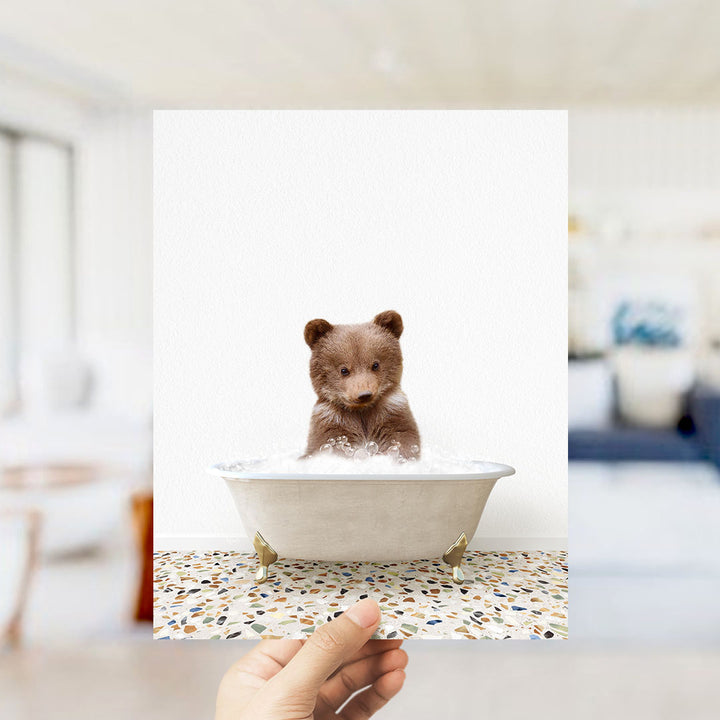 A small brown bear sitting in a white bathtub filled with bubbles, with a hand holding the image up to the camera.