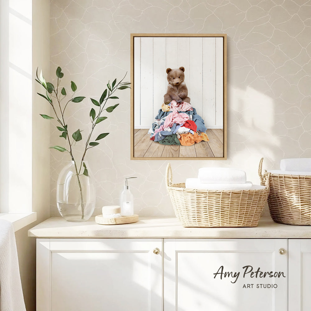 a framed photograph of a small brown bear sitting on a pile of clothes, hanging on a wall above a white cabinet.