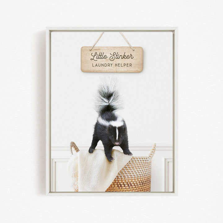 A black and white skunk standing on a woven basket, with a wooden sign above that says "Little Stinker Laundry Helper".