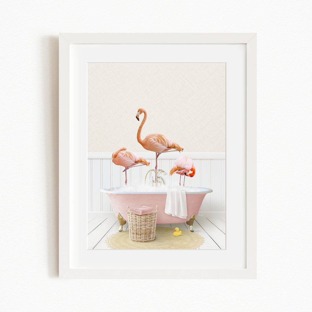 A framed image depicting three pink flamingos standing in a pink bathtub, with a basket of bath toys and a yellow rubber duck nearby.