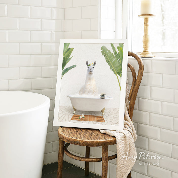 A framed image of a white llama sitting in a bathtub, surrounded by tropical plants, is displayed on a wooden chair in a bathroom setting.