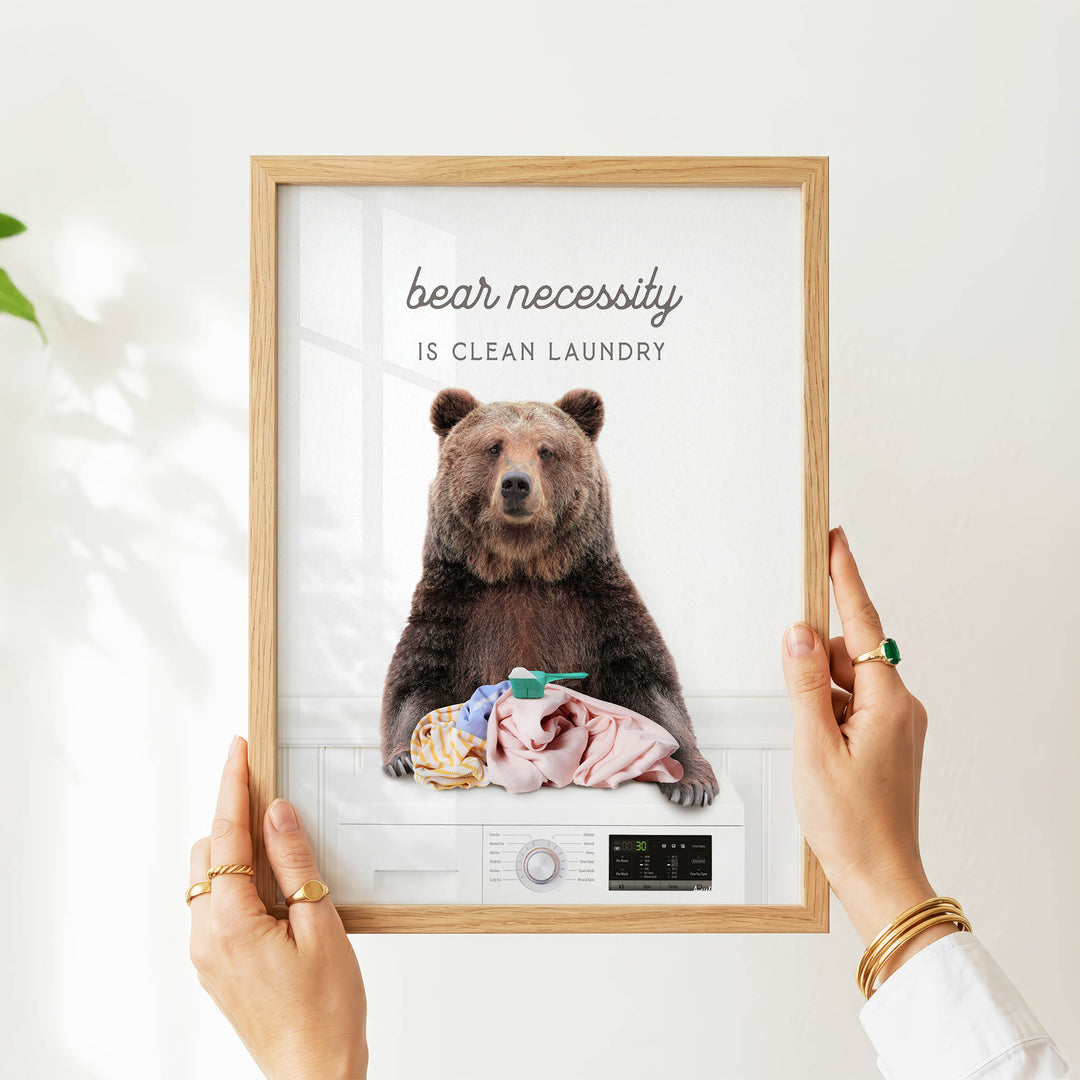A person is holding up a framed image of a bear sitting on a washing machine, with the text "bear necessity is clean laundry" displayed above the bear.