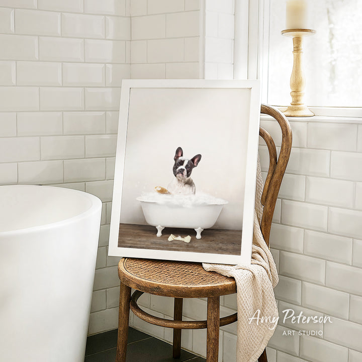 A framed photograph of a dog sitting in a bathtub, with a towel draped over the back of a wooden chair in a bathroom setting.