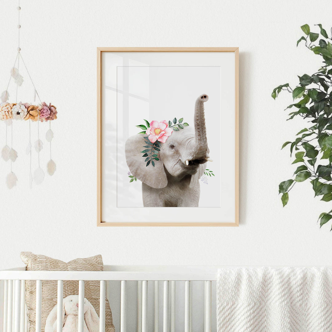 A framed picture of an elephant with a floral headpiece hangs on a white wall, accompanied by a hanging mobile and a potted plant in a nursery setting.