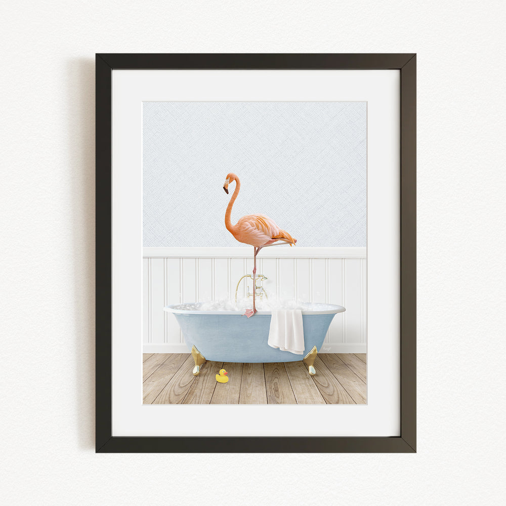 A framed image depicting a pink flamingo standing on the edge of a blue bathtub filled with water, with a white towel and yellow rubber duckies nearby.