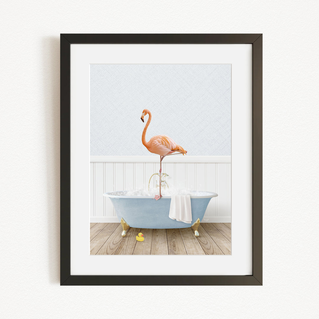 A framed image depicting a pink flamingo standing on the edge of a blue bathtub filled with water, with a white towel and yellow rubber duckies nearby.
