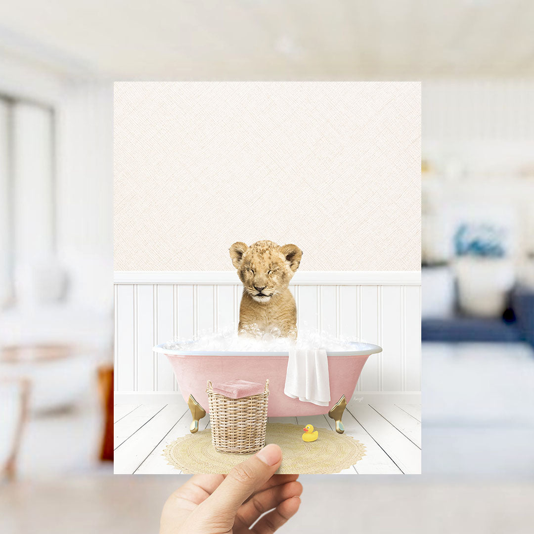 A hand holds up a framed image depicting a young lion cub sitting in a pink bathtub filled with bubbles, surrounded by a yellow rubber duck and a white towel.
