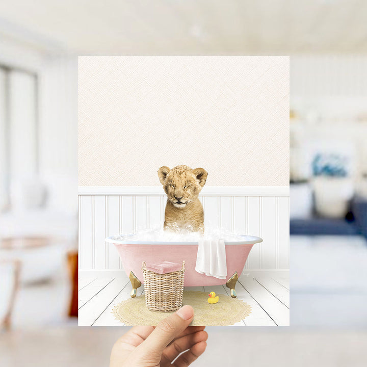 A hand holds up a framed image depicting a young lion cub sitting in a pink bathtub filled with bubbles, surrounded by a yellow rubber duck and a white towel.