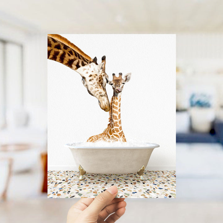A hand holds up a framed photograph of two giraffes, one adult and one juvenile, standing in a bathtub filled with water.