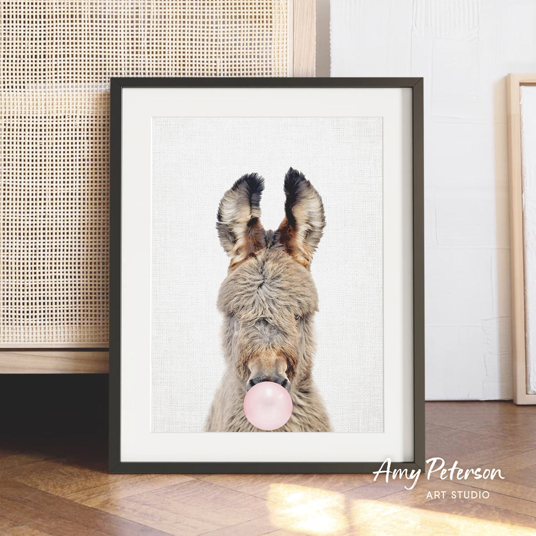 A framed photograph of a donkey blowing a bubble gum bubble, with a wooden floor and a woven basket in the background.