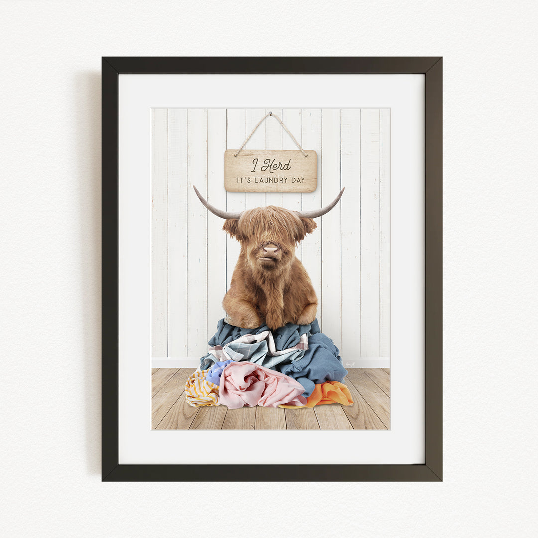 A framed image of a Highland cow with large horns sitting on a pile of clothes, with a sign above it that says "It's laundry day".