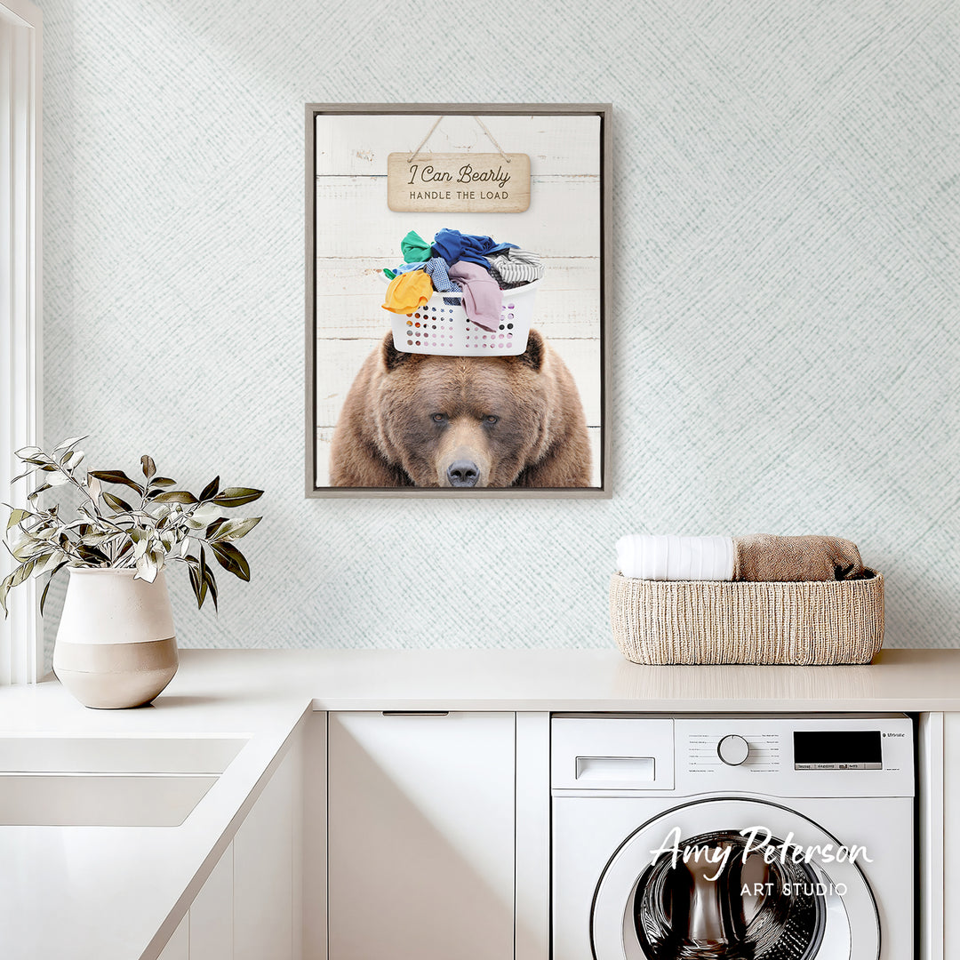 a framed photo of a bear wearing a basket on its head, with the caption "I can bear the load" and the text "Handles the load" below the bear. The bear is sitting on a pile of laundry, and there is a basket of clothes nearby. The backdrop is a white wall with a patterned design.