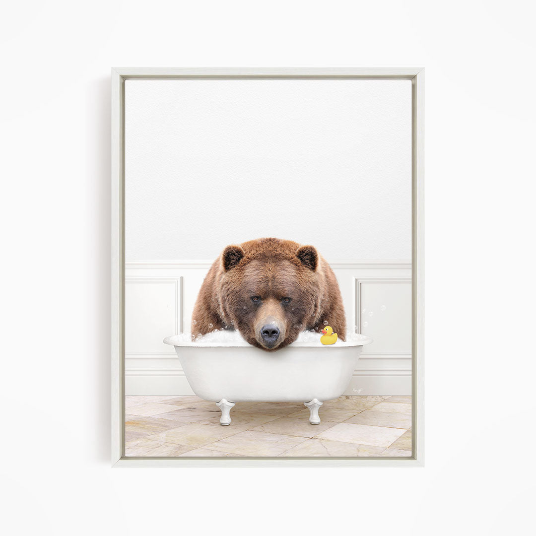 A brown bear is sitting in a white bathtub, with a yellow rubber duck floating in the water.