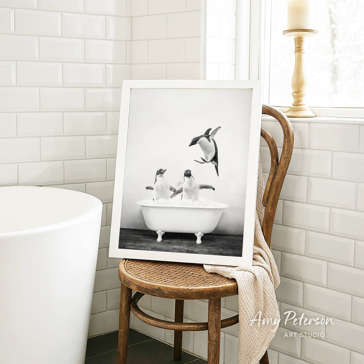 A framed black and white photograph of two penguins sitting in a bathtub, with one penguin jumping out of the tub. The photograph is displayed on a wooden chair in a bathroom setting.