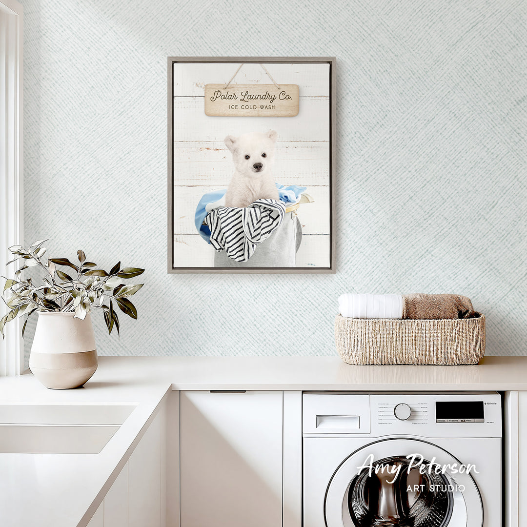 a framed picture of a white dog sitting in a bucket, with a sign above it that reads "Polar Laundry Co. Ice Cold Wash". The scene is set in a white-themed room with a white countertop, a potted plant, and a basket of towels.