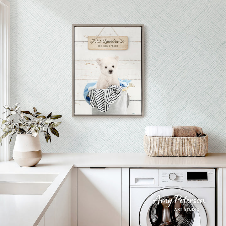 a framed picture of a white dog sitting in a bucket, with a sign above it that reads "Polar Laundry Co. Ice Cold Wash". The scene is set in a white-themed room with a white countertop, a potted plant, and a basket of towels.