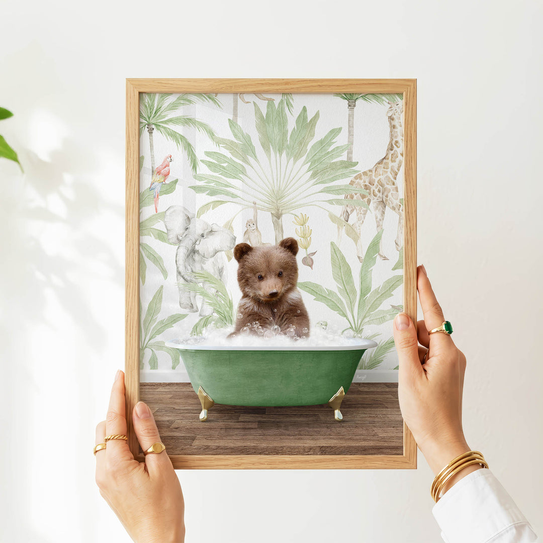 A person's hands are holding a framed picture of a small brown bear sitting in a green bathtub, surrounded by tropical foliage.