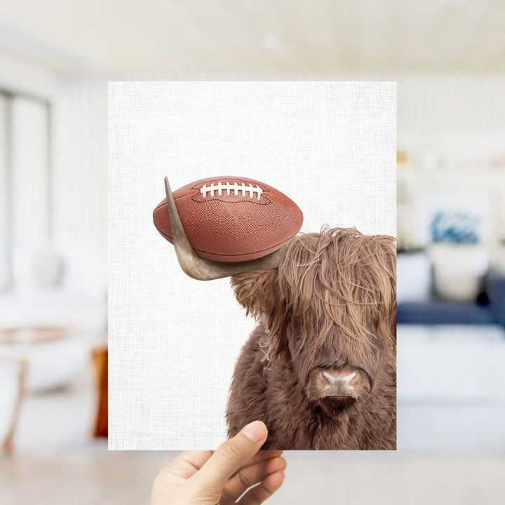 A hand holds up a framed image of a Highland cow with a football on its head, creating a playful and whimsical scene.