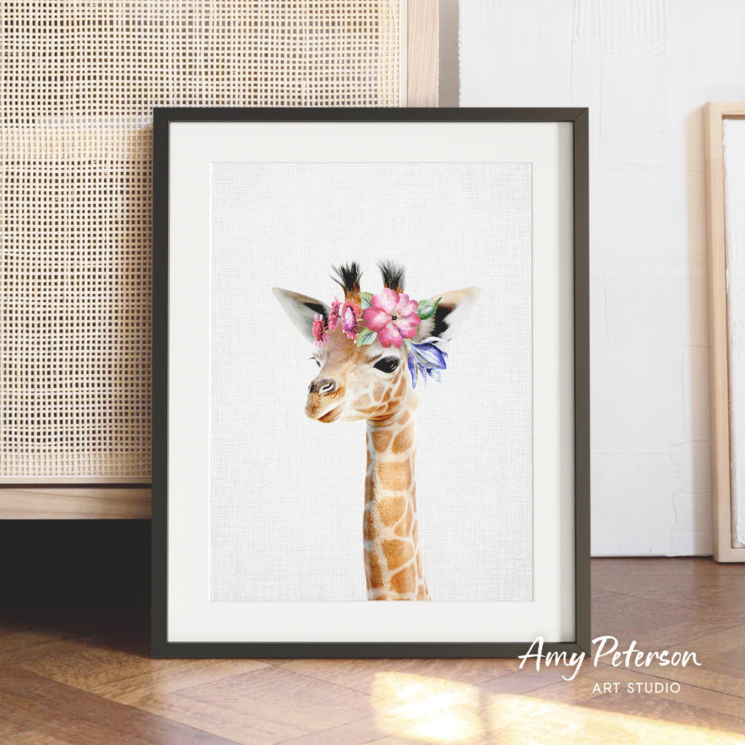 A framed print of a giraffe wearing a flower crown is displayed on a wooden floor against a white wall.