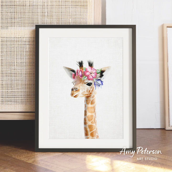 A framed print of a giraffe wearing a flower crown is displayed on a wooden floor against a white wall.