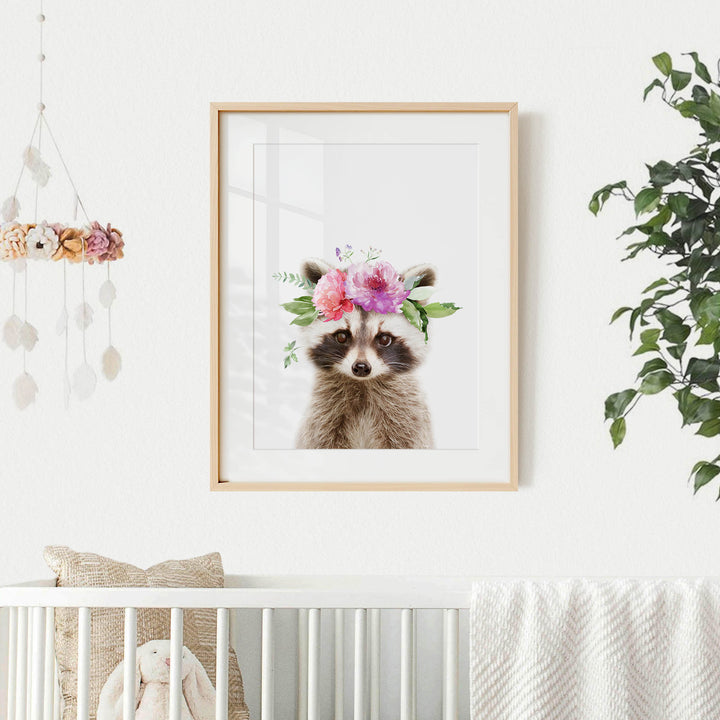 A framed picture of a raccoon wearing a floral crown is displayed on a white wall, with a hanging mobile and a plant in the background.
