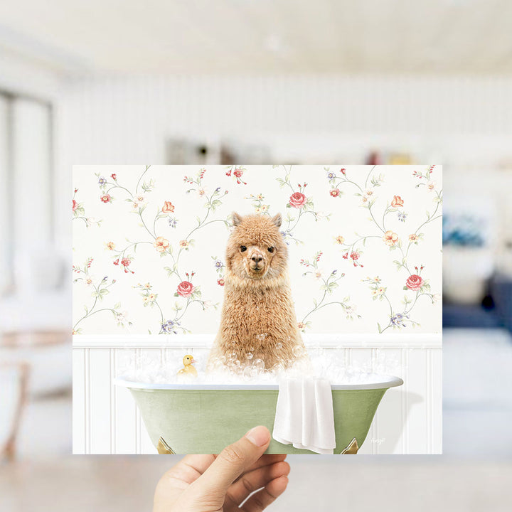 A hand holding a framed image of a brown alpaca sitting in a green bathtub, surrounded by a floral pattern.