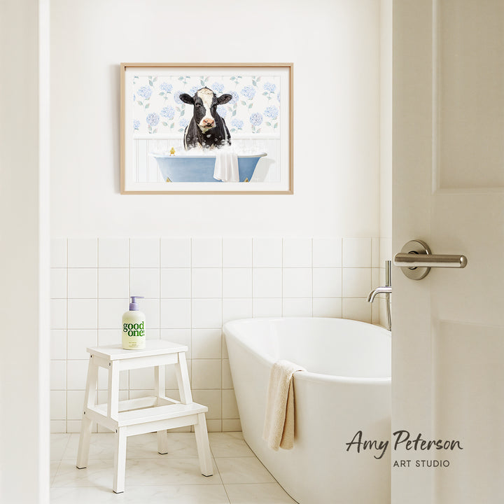 A framed painting of a cow in a bathtub hangs on a white tiled wall above a white bathtub in a bathroom.
